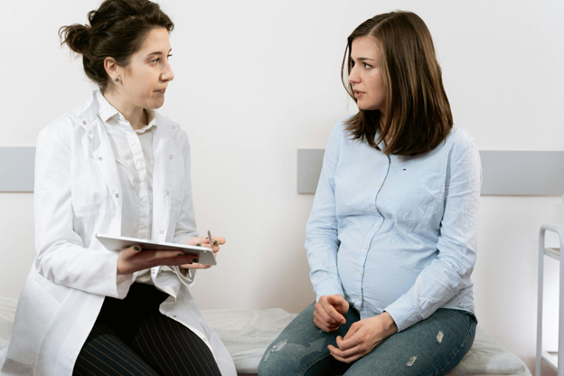 A female doctor in a white coat holds a tablet while discussing medical information with a pregnant patient sitting on a hospital bed. The patient listens attentively, and the setting is a bright and clean medical examination room.
